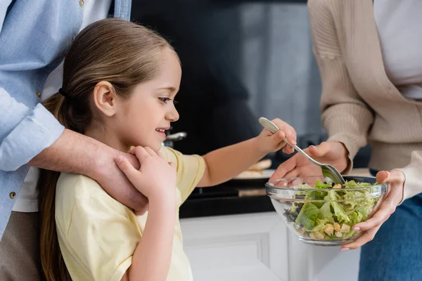 Joyeuse fille mélangeant salade de légumes frais dans un bol près des parents dans la cuisine — Photo de stock