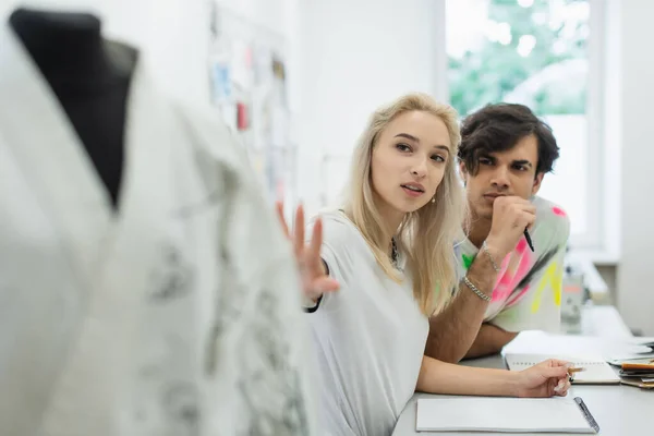 Fashion designer pointing at blurred mannequin near colleague in atelier — Stock Photo