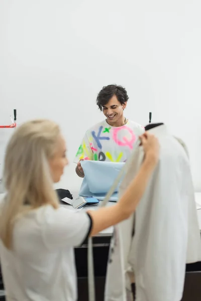 Designer sorridente guardando campione di tessuto vicino collega misurare vestiti su manichino in primo piano sfocato — Foto stock
