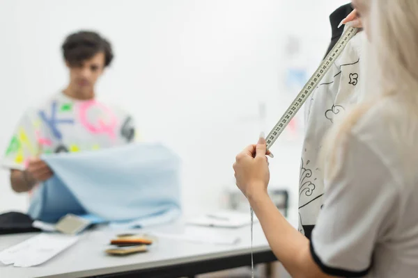 Designer de moda roupas de medição no manequim perto de colega borrado segurando amostra de tecido — Fotografia de Stock