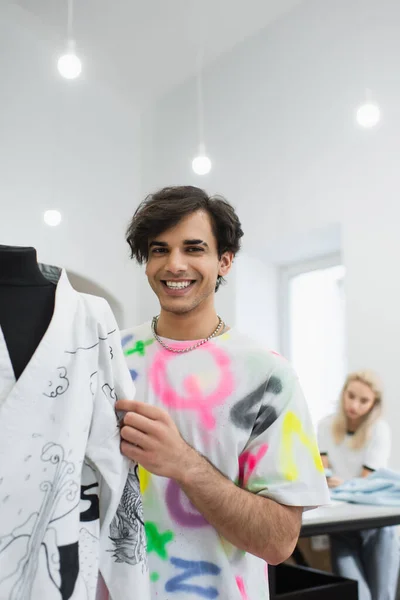 Designer alla moda sorridente alla fotocamera vicino kimono sul manichino e collega che lavora su sfondo sfocato — Foto stock