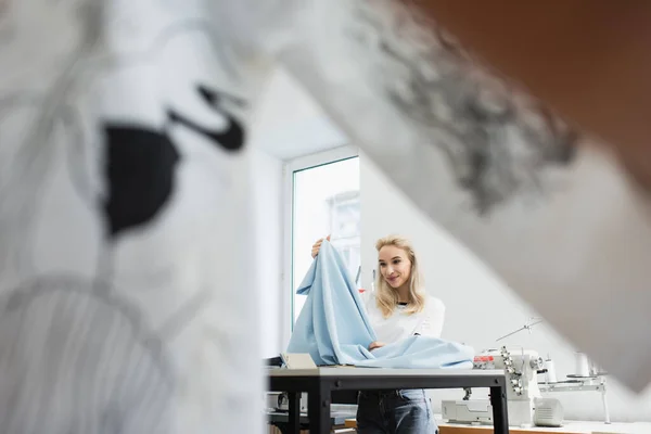 Happy fashion designer sitting at workplace and looking at fabric sample, blurred foreground — Stock Photo