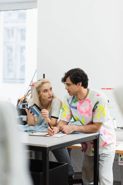 Jeune designer tenant une palette de couleurs tout en parlant à un collègue dans un magasin de tailleur, au premier plan flou — Photo de stock