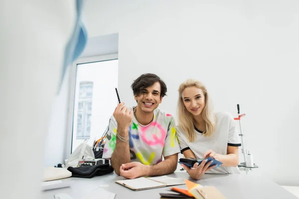 Smiling fashion designers looking at camera while choosing colors from palette on blurred foreground — Stock Photo