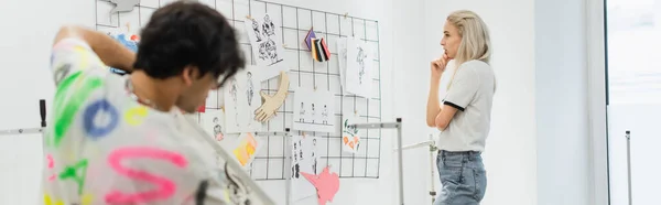 Thoughtful fashion designer looking at sketches on wall near blurred colleague, banner — Stock Photo