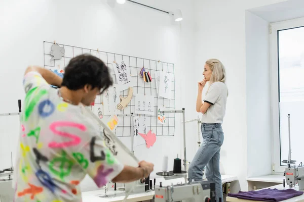Blonde fashion designer thinking near sketches on wall and blurred colleague with measuring tape — Stock Photo