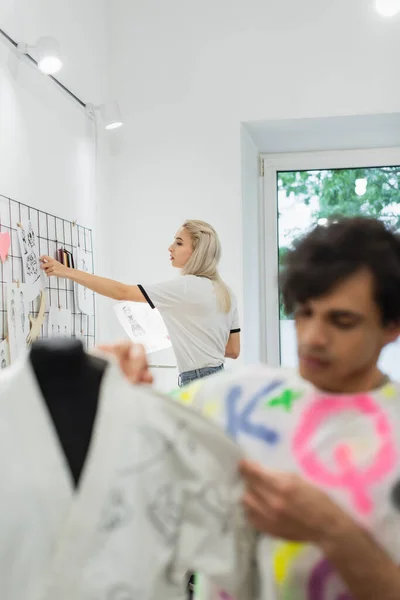 Junge Designerin befestigt Zeichnungen an Wand in der Nähe von Kollegen, die an verschwommenem Vordergrund arbeiten — Stockfoto