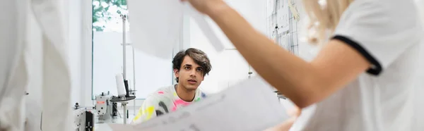 Junge Modedesignerin blickt verschwommenen Kollegen in Schneiderei an, Banner — Stockfoto