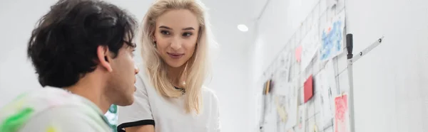 Joven diseñador hablando con rubia colega cerca de dibujos en la pared, pancarta - foto de stock