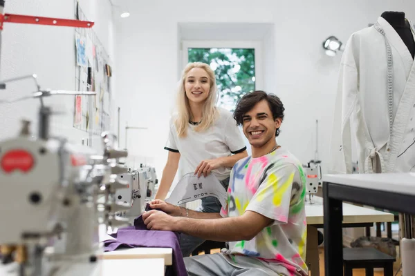 Happy tailor and fashion designer looking at camera near sewing machines in atelier — Stock Photo
