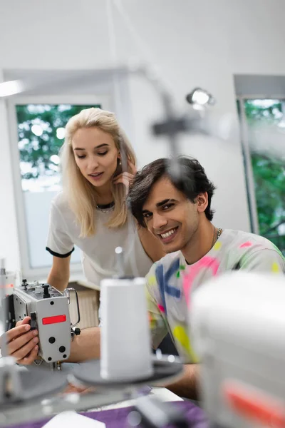 Feliz sastre mirando a la cámara mientras regula la máquina de coser cerca de diseñador de moda - foto de stock