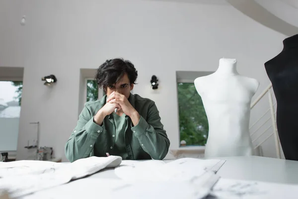 Alfaiate pensativo sentado perto de padrões borrados de costura e tecido no atelier — Fotografia de Stock