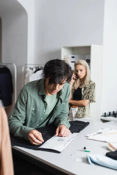 Junge Schneiderin arbeitet mit Schnittmuster und Tuch in der Nähe des Designers und spricht auf Smartphone auf verschwommenem Hintergrund — Stockfoto