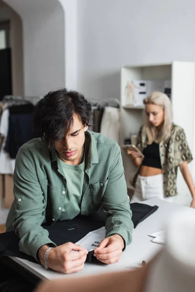 Jeune tailleur travaillant avec motif de couture et tissu près du concepteur en utilisant smartphone sur fond flou — Photo de stock