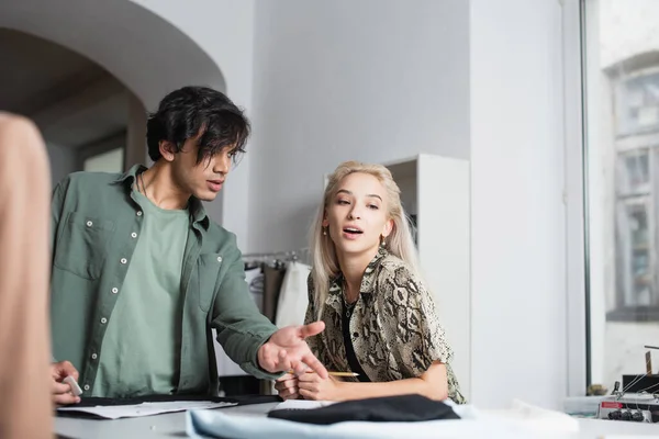 Diseñador rubia hablando con sastre apuntando a tejido en primer plano borrosa - foto de stock