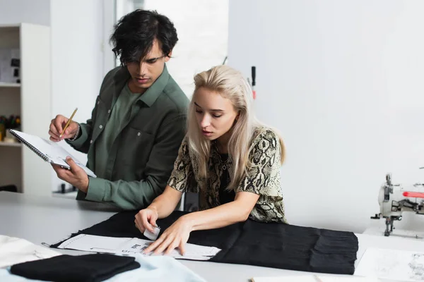 Estiloso designer de escrita em notebook perto de tecido de corte costureiro no atelier — Fotografia de Stock