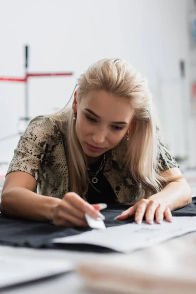 Young dressmaker cutting tissue near sewing pattern on blurred foreground — Stock Photo