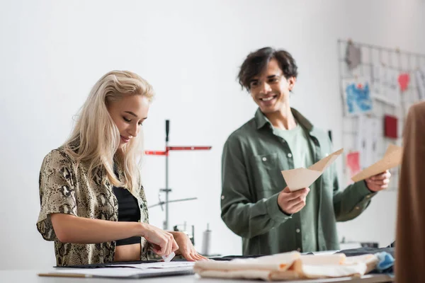 Cheerful designer with sewing patterns looking at dressmaker cutting tissue — Stock Photo