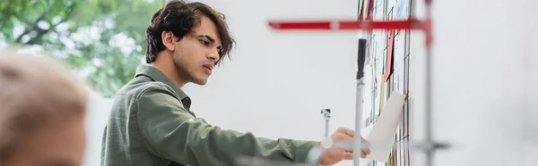 Joven diseñador de moda mirando bocetos en la pared cerca borrosa colega, pancarta - foto de stock