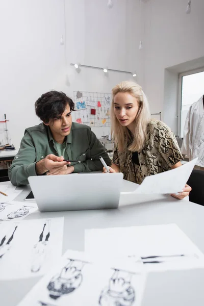 Young designer pointing at laptop near blonde colleague and blurred sketches — Stock Photo