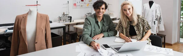 Jóvenes diseñadores sonriendo a la cámara cerca de gadgets y bocetos en el lugar de trabajo, pancarta - foto de stock