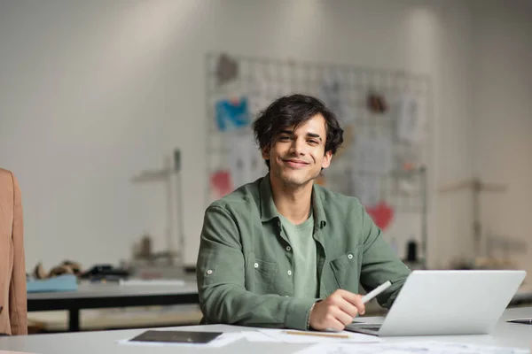 Diseñador alegre sonriendo a la cámara cerca del portátil en el taller de moda - foto de stock