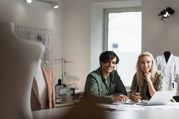 Glückliche Designer lächeln in die Kamera, während sie in der Modewerkstatt auf verschwommenem Vordergrund arbeiten — Stockfoto