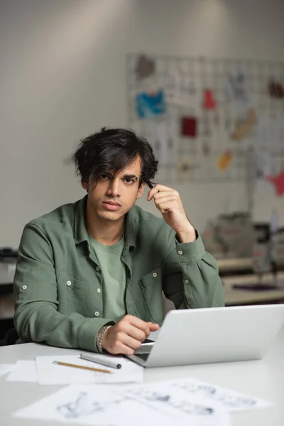 Elegante diseñador de moda mirando a la cámara mientras está sentado cerca de la computadora portátil en atelier - foto de stock