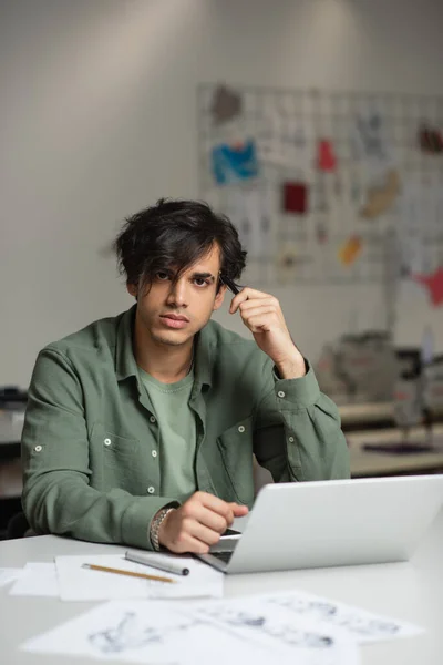 Styliste de mode élégant regardant la caméra tout en étant assis près de l'ordinateur portable dans l'atelier — Photo de stock