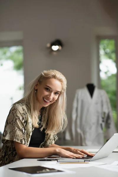 Cheerful fashion designer looking at camera while typing on laptop in atelier — Stock Photo