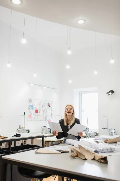 Emocionado diseñador de moda riendo mientras está sentado en el lugar de trabajo en la amplia sastrería - foto de stock