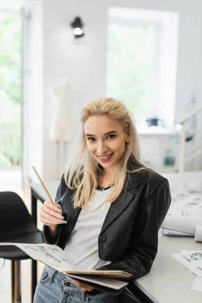Cheerful fashion designer with notebook, pencil and drawing looking at camera in atelier — Stock Photo