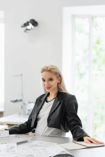 Smiling fashion designer looking at camera near drawings and sewing patterns — Stock Photo