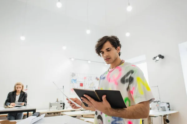 Modedesigner steht mit Notizbuch und Papier neben Mitarbeiter auf verschwommenem Hintergrund — Stockfoto