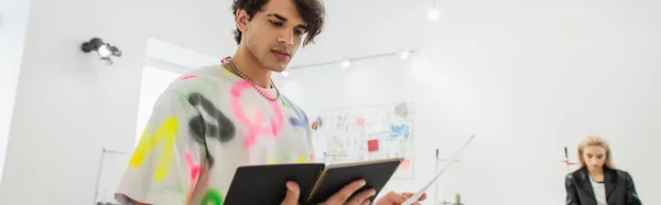 Trendige Modedesignerin blickt auf Notizbuch neben verschwommenem Kollegen im Atelier, Banner — Stockfoto