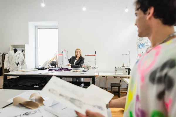 Designer de moda sorridente mostrando padrão de costura ao colega em primeiro plano borrado — Fotografia de Stock