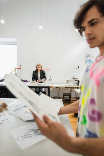 Difuminado diseñador de moda mirando dibujo cerca colega trabajando en segundo plano - foto de stock