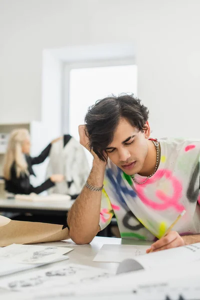 Tired fashion designer sitting with closed eyes near sketches and colleague on blurred background — Stock Photo