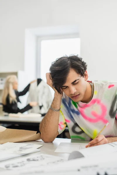 Diseñador de moda cansado sentado con los ojos cerrados cerca de bocetos y colega sobre fondo borroso - foto de stock