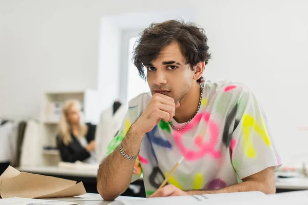 Stylischer Modedesigner blickt in die Kamera, während er am Arbeitsplatz in der Nähe eines Kollegen auf verschwommenem Hintergrund sitzt — Stockfoto