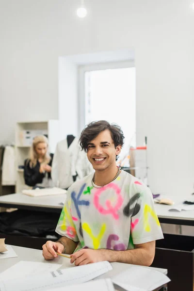 Fröhliche Modedesignerin blickt in Kamera neben Kollegin, die an verschwommenem Hintergrund arbeitet — Stockfoto