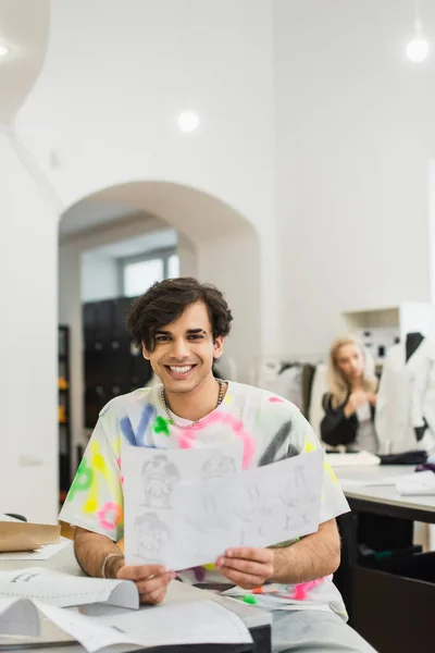 Fashion designer with sketches smiling at camera near blurred colleague — Stock Photo