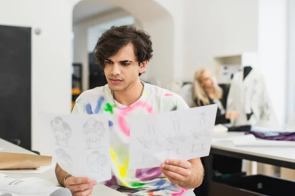 Estilista elegante olhando para desenhos perto de colega de trabalho no fundo borrado — Fotografia de Stock