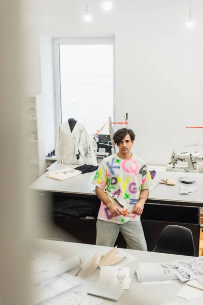 Vue grand angle du créateur de mode à la mode regardant la caméra près du bureau avec des motifs de couture, avant-plan flou — Photo de stock