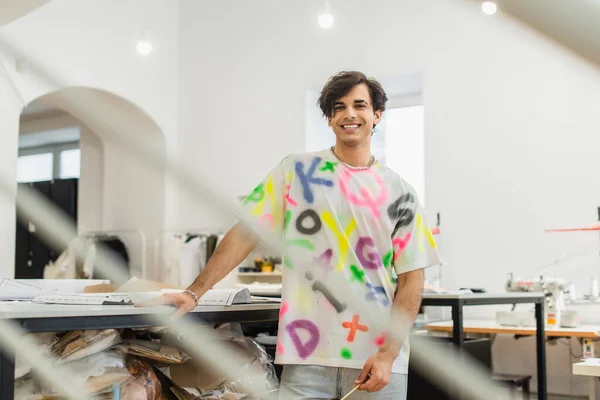 Diseñador de moda feliz sonriendo a la cámara cerca de los patrones de costura en el escritorio, borrosa primer plano - foto de stock