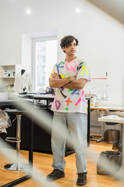 Joven y serio diseñador de moda de pie con los brazos cruzados en el taller - foto de stock