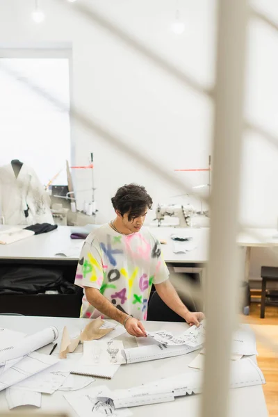 Fashionable designer looking at sketches in atelier on blurred foreground — Stock Photo