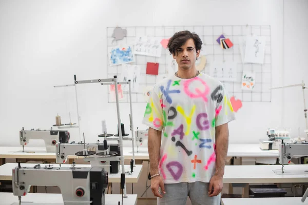 Stylish fashion designer looking at camera while standing near sewing machines in atelier — Stock Photo