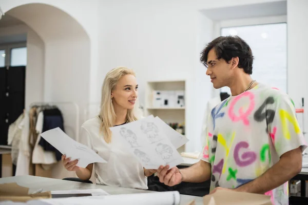 Estilistas elegantes segurando esboços e olhando uns para os outros enquanto conversam no atelier — Fotografia de Stock