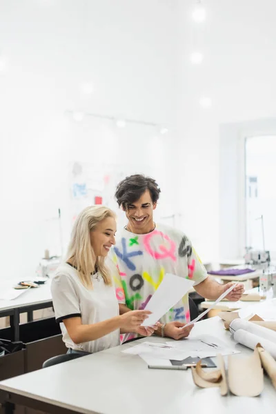 Diseñadores de moda felices sosteniendo papeles mientras trabajan en sastrería - foto de stock